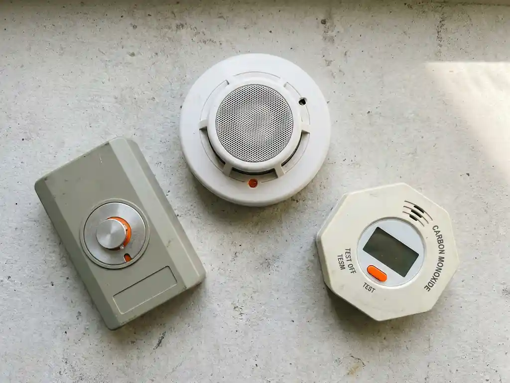 Drie brandmelders op betonnen ondergrond: rookmelder, hittemelder en CO-detector in grijze en witte tinten met oranje accenten.